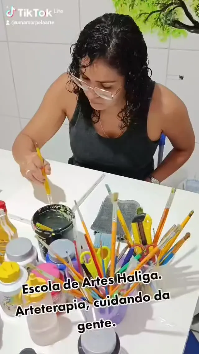 Mulher concentrada pintando, com potes de pincéis e tintas na mesa.