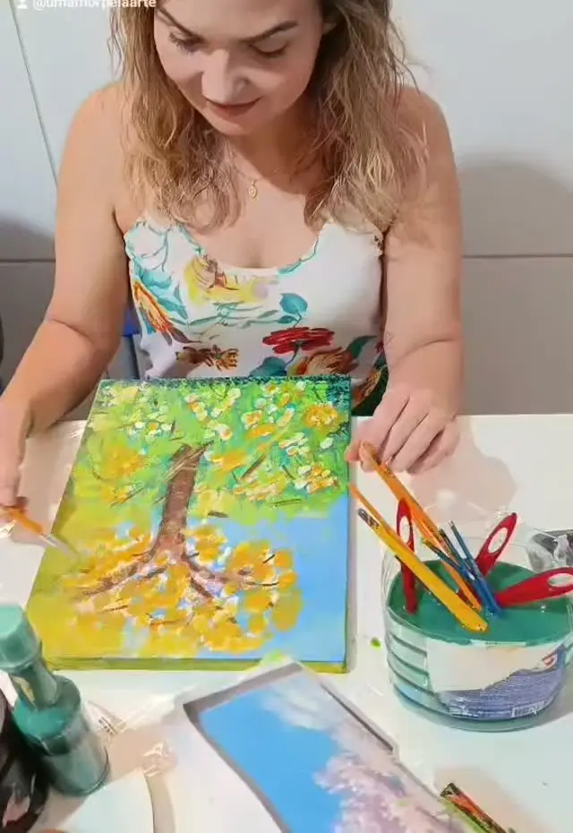 Mulher pintando uma tela com uma árvore de folhas amarelas sobre fundo verde.