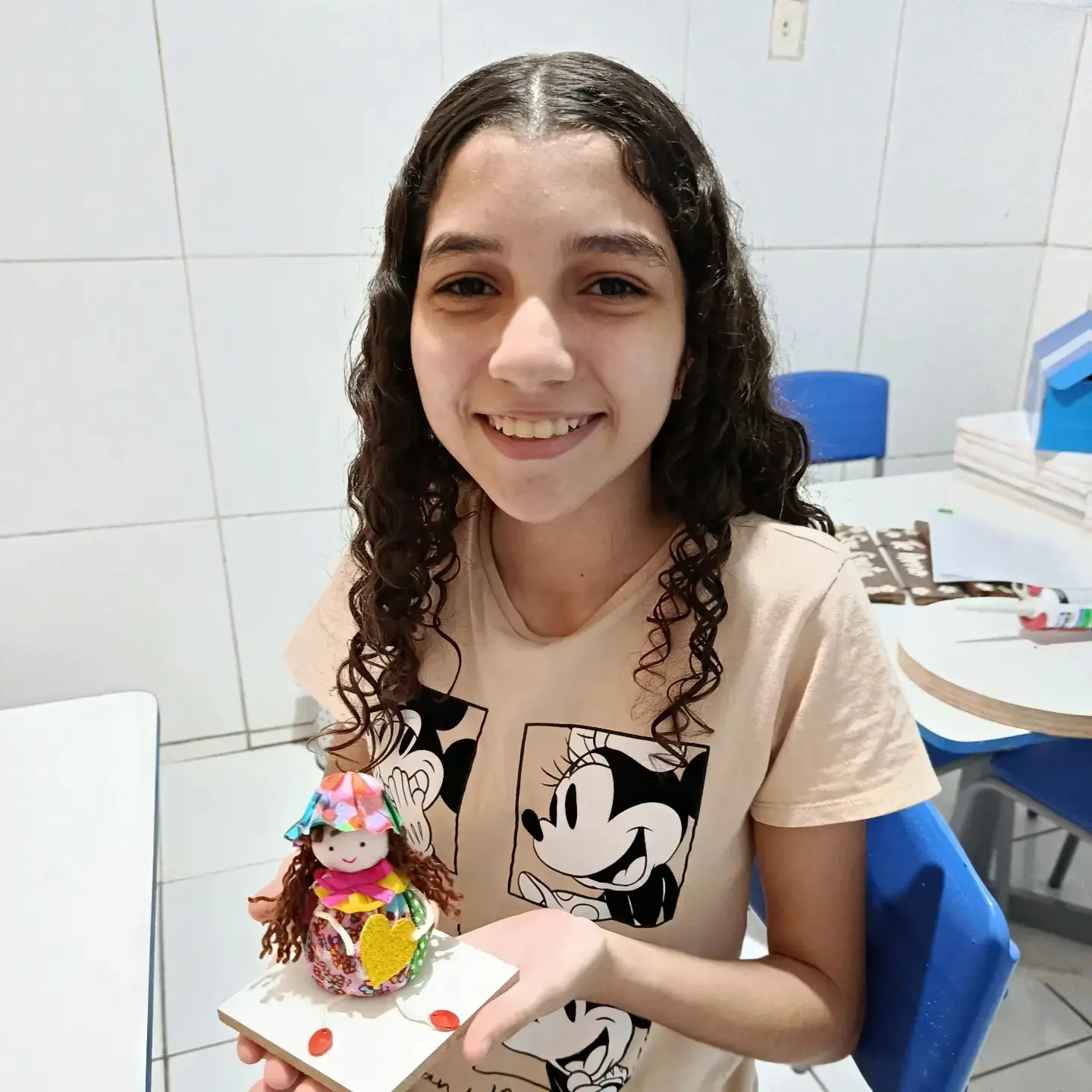 Menina sorrindo e segurando uma bonequinha de pano que ela mesma fez.