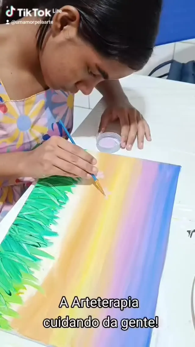 Mão pintando um céu em degradê de amarelo, rosa e lilás sobre a grama verde.
