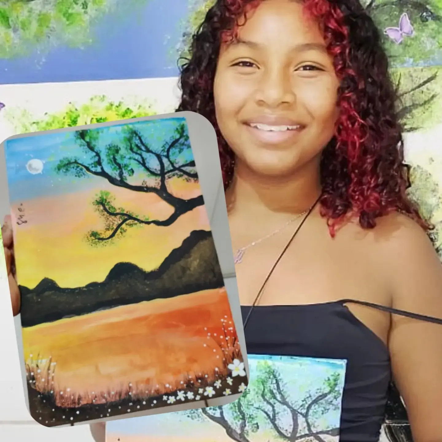 Menina de cabelo cacheado vermelho sorrindo e segurando a tela de um pôr do sol com uma árvore.