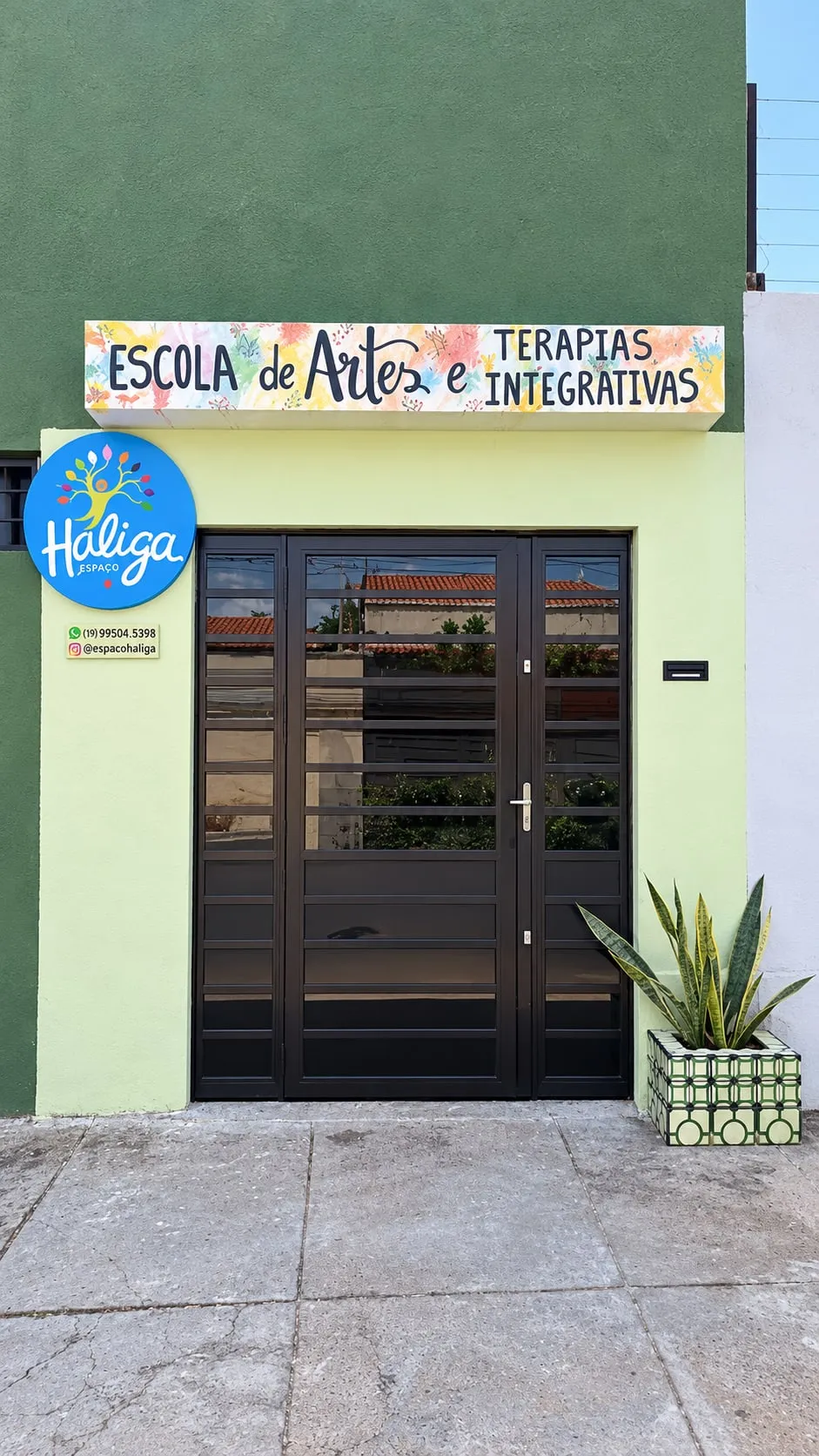 Fachada verde da Haliga, com o letreiro colorido “Escola de Artes e Terapias Integrativas”, o selo azul ao lado da porta e um vaso de espada-de-são-jorge.