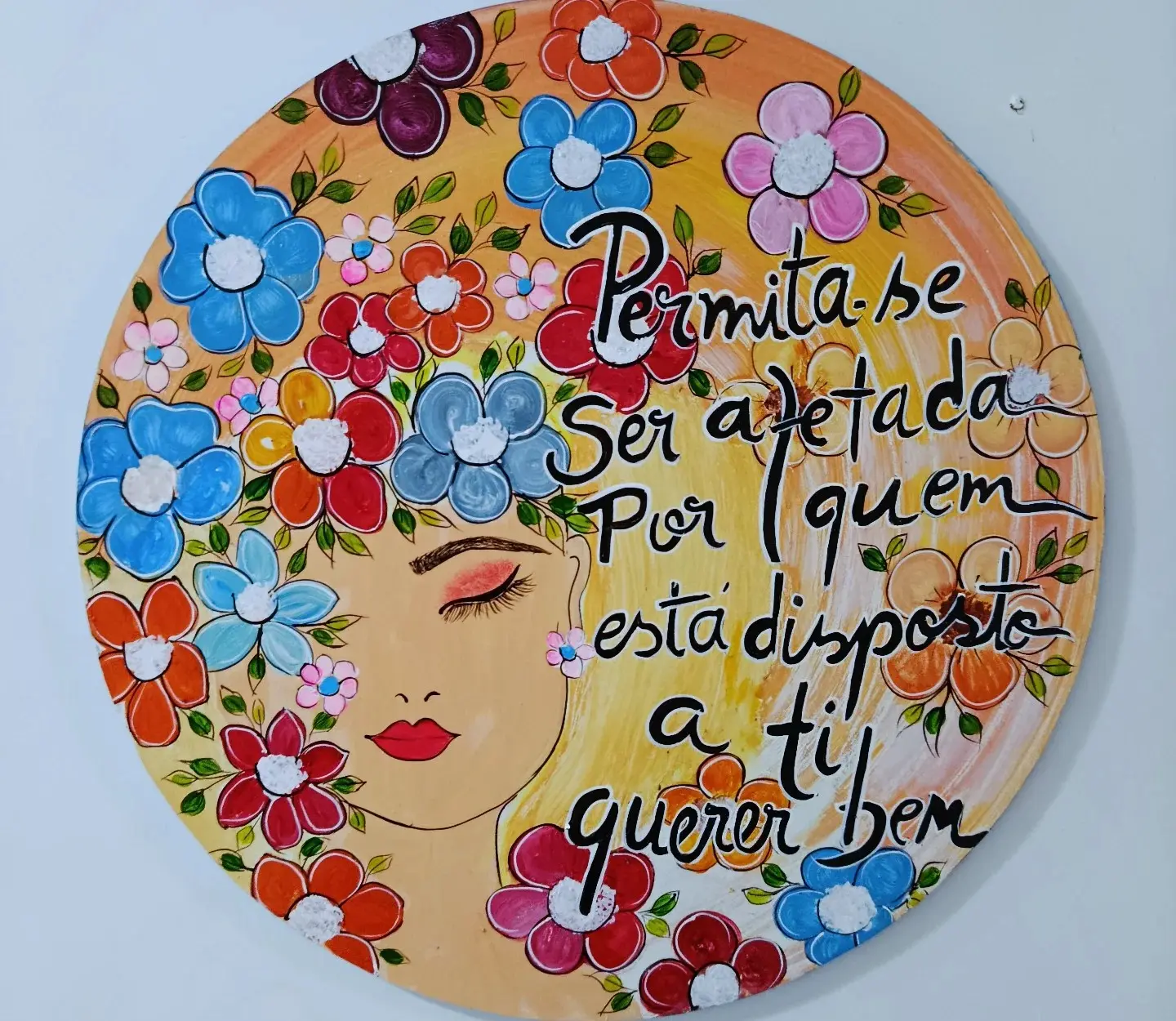 Mandala redonda com um rosto de mulher cercado de flores coloridas e uma frase pintada à mão.