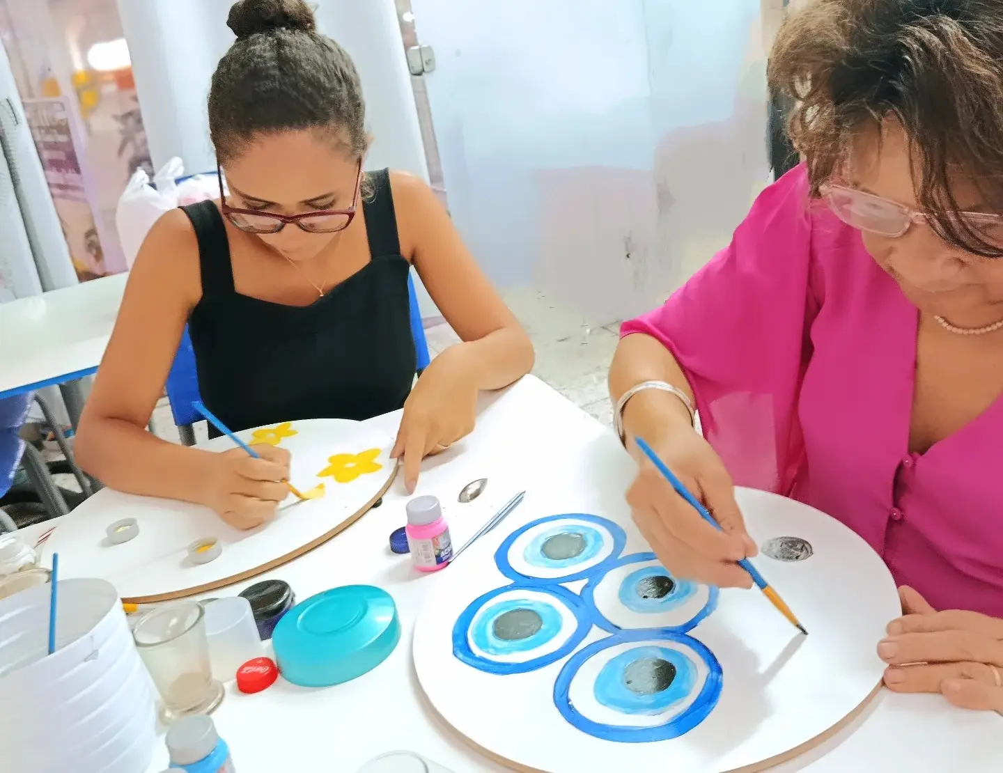 Duas mulheres pintando mandalas em círculos brancos de madeira, com tintas azuis na mesa.