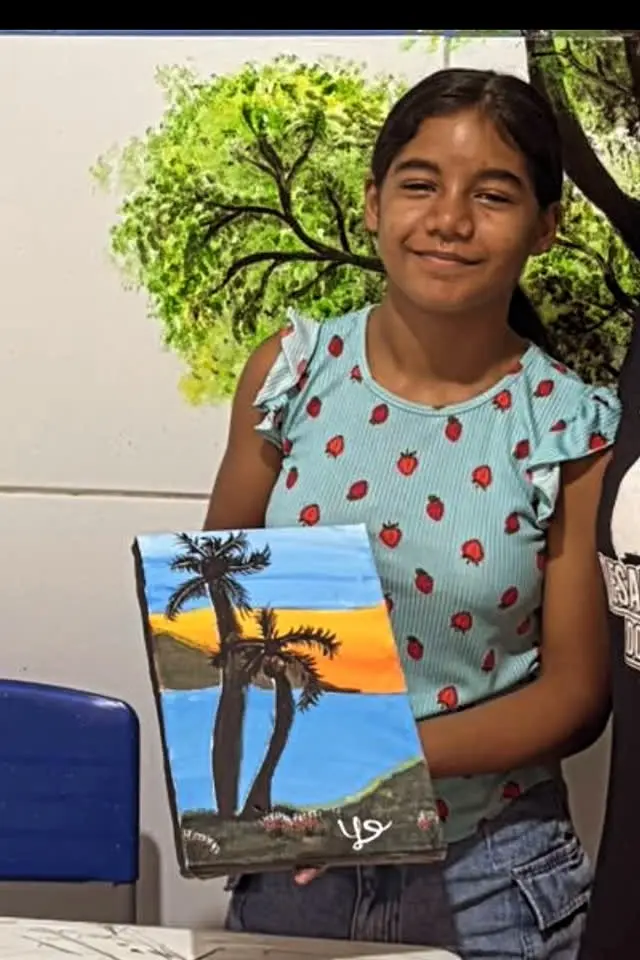 Menina sorrindo e mostrando uma tela com palmeiras contra o céu laranja do fim de tarde.