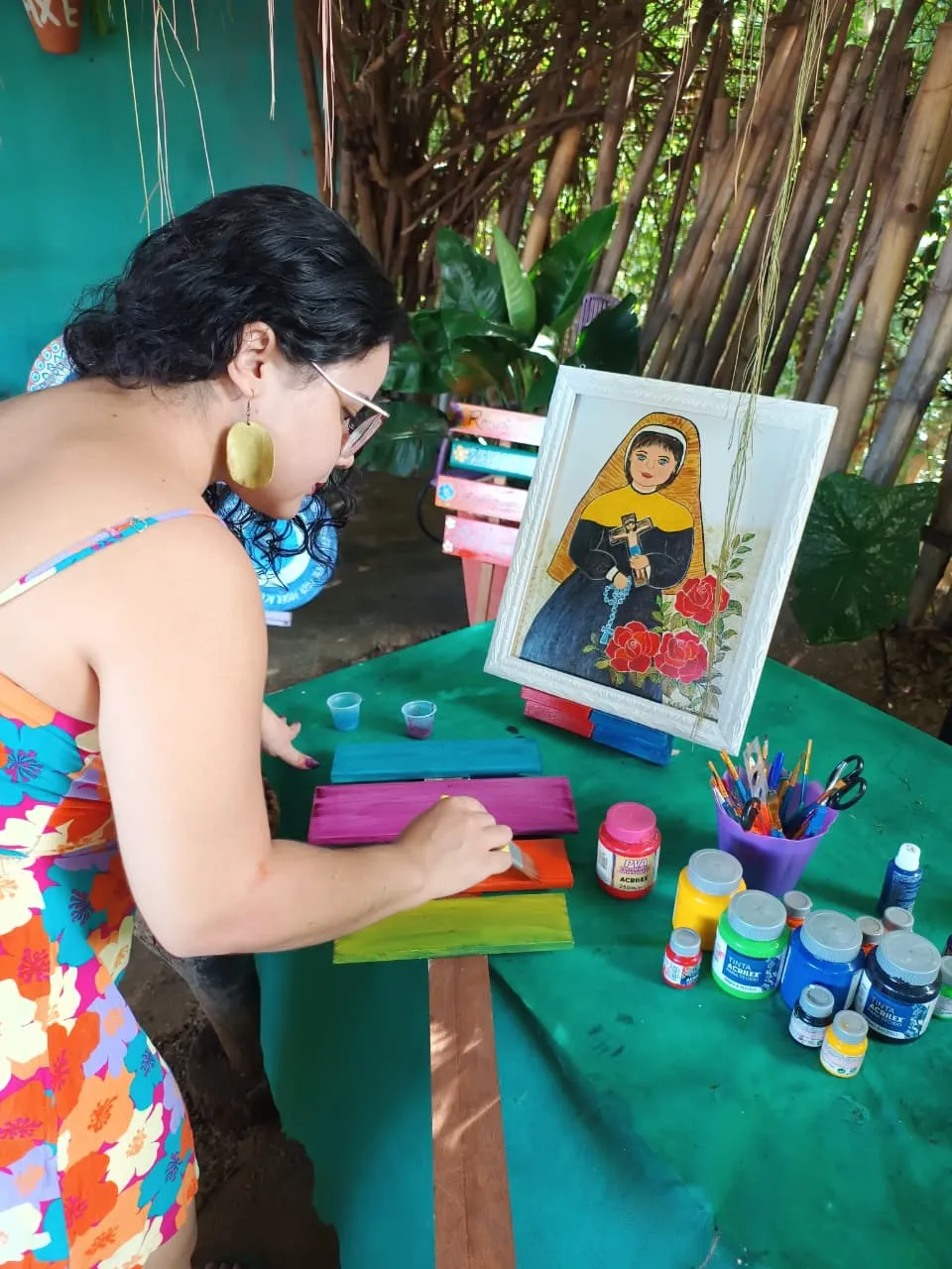 Aluna pintando plaquinhas coloridas no quintal, com uma tela de Nossa Senhora ao fundo.