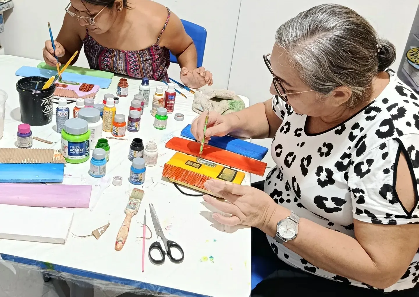 Duas alunas pintando plaquinhas de madeira lado a lado, com tintas e pincéis espalhados pela mesa.
